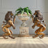 A pair of vintage ornate bronze lion plaques facing each other on either side of a distressed white stone urn pedestal with a green fern arrangement, set against a paneled gray wall and patterned beige carpet.