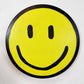 Smiley 1 - By Drew Outsider art painting, A round yellow smiley face sticker or decal with a black border, black oval eyes, and a black curved smile on a plain white background.