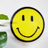A round yellow smiley face painting by Drew outsider artist with a black border, black eyes, and a black curved smile, photographed on a white background with green leaves at the left edge.