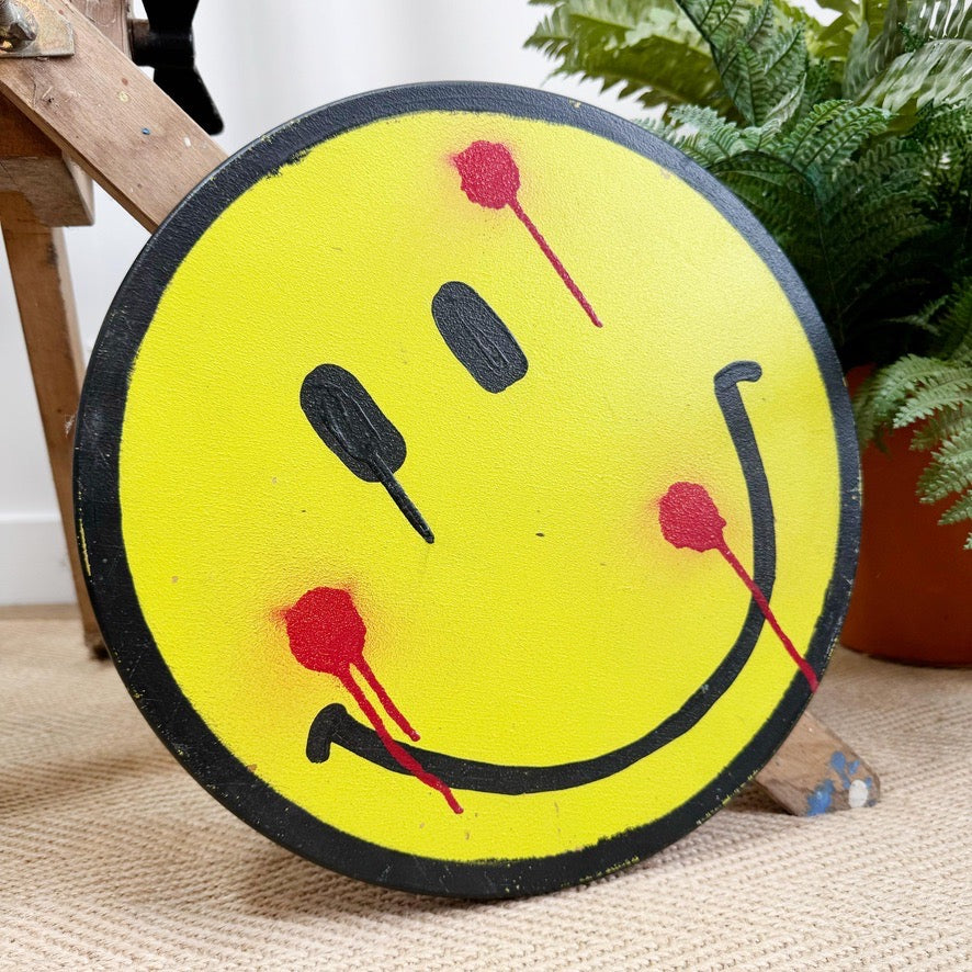 A round yellow sign featuring a black smiley face with red splatter details, by Drew outsider artist, photographed indoors on a woven rug with a plant and wooden furniture in the background.