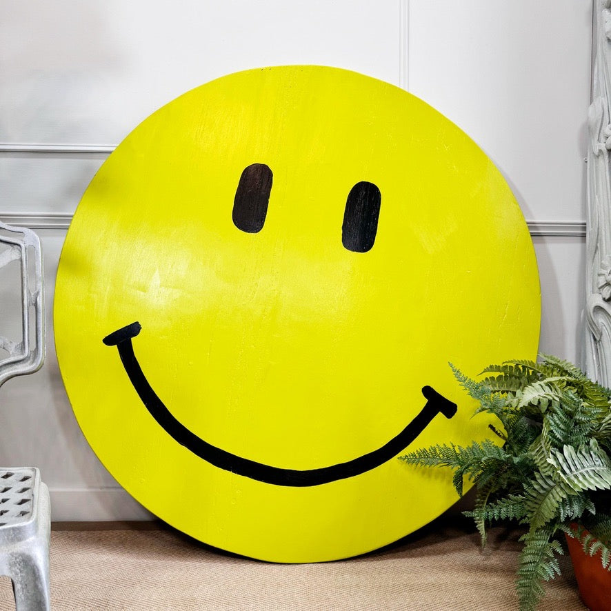 A large yellow smiley face painting by outsider artist Drew, with black eyes and a wide black smile, leaning against a white paneled wall next to a green potted fern.