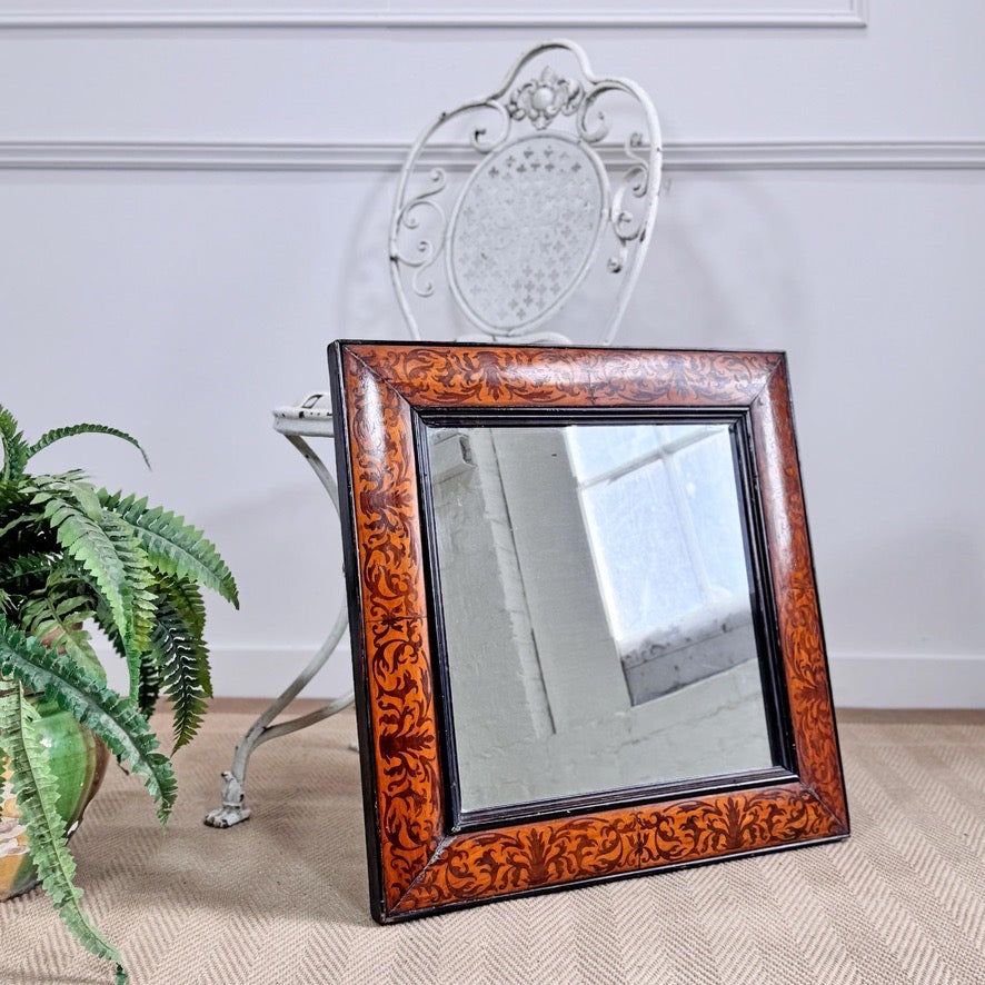 A square William and Mary Walnut and Seaweed Marquetry Mirror Circa 1690 with an ornate reddish-brown marquetry frame, leaning against a white metal chair in a styled interior setting with a potted fern on the left.