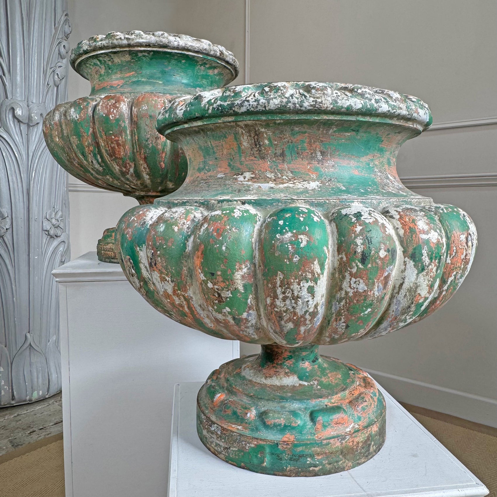 Two large distressed painted iron Alfred Corneau  urns with a peeling green and white finish, displayed on white pedestals in an indoor setting.