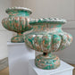 Two distressed Alfred Corneau teal-green antique urn planters with a weathered, peeling paint finish, displayed on white pedestals in an indoor setting.