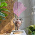 A decorative pink fan coral sculpture displayed in an ornate antique wooden and copper repousse altar urn on a white pedestal, set against a patterned gold wallpaper and green plants.