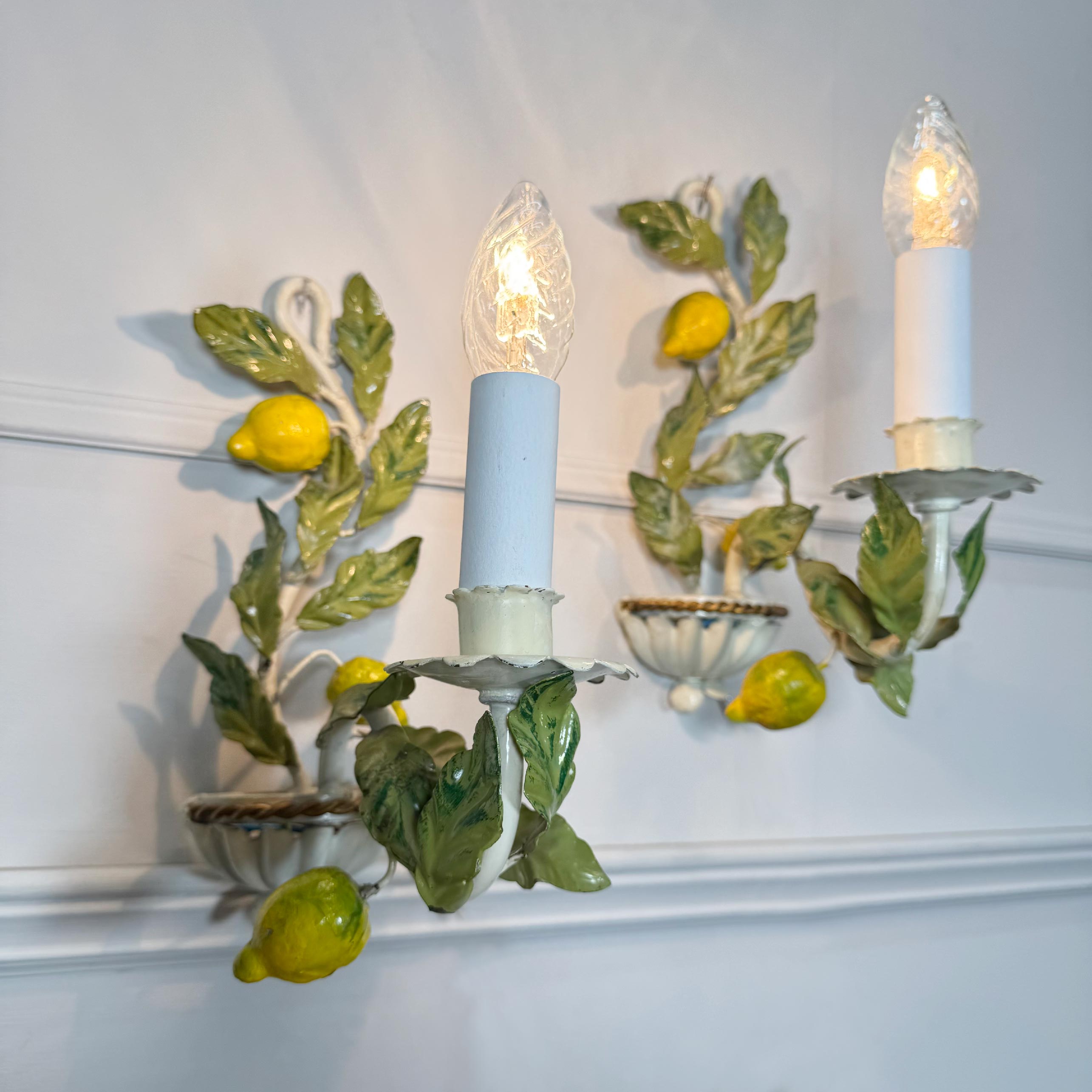 A decorative Italian vintage Lemons Toleware sconce with three candle-style lights, featuring sculpted green leaves, yellow lemons, and white ceramic bowls against a white paneled wall.