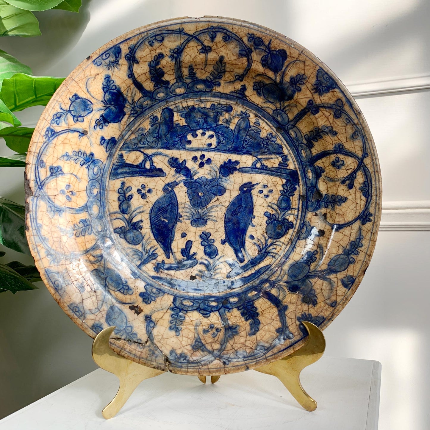 An 16th Century Safavid Pottery Dish plate with a blue and beige floral design, displayed on a gold-colored stand against a white wall with soft shadows.