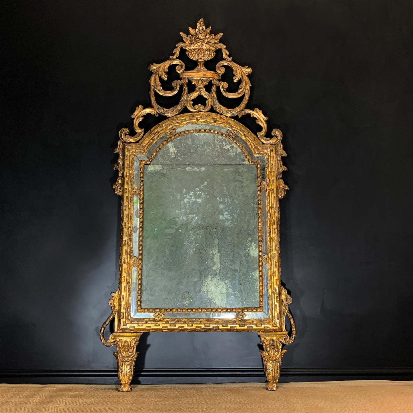 An ornate Early 18th Century Giltwood Antique Italian Mirror with a gilded gold wooden  frame and a green-tinted glass panel, set against a dark background.