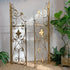 Two ornate gold-toned metal iron gates with scrollwork and fleur-de-lis motifs, displayed indoors against a white paneled wall with potted green plants around them.