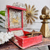 An ornate open red antique wooden box with carved silver-toned interior panels and gold-toned decorative pieces inside, displayed on a dark wooden table with coral, a leafy plant, and a gold wall sun in the background.