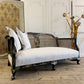 An antique settee with a black and gold chinoiserie lacquered frame, woven cane backrest, and off-white upholstered seat, styled with three white pillows in a rustic room setting.
