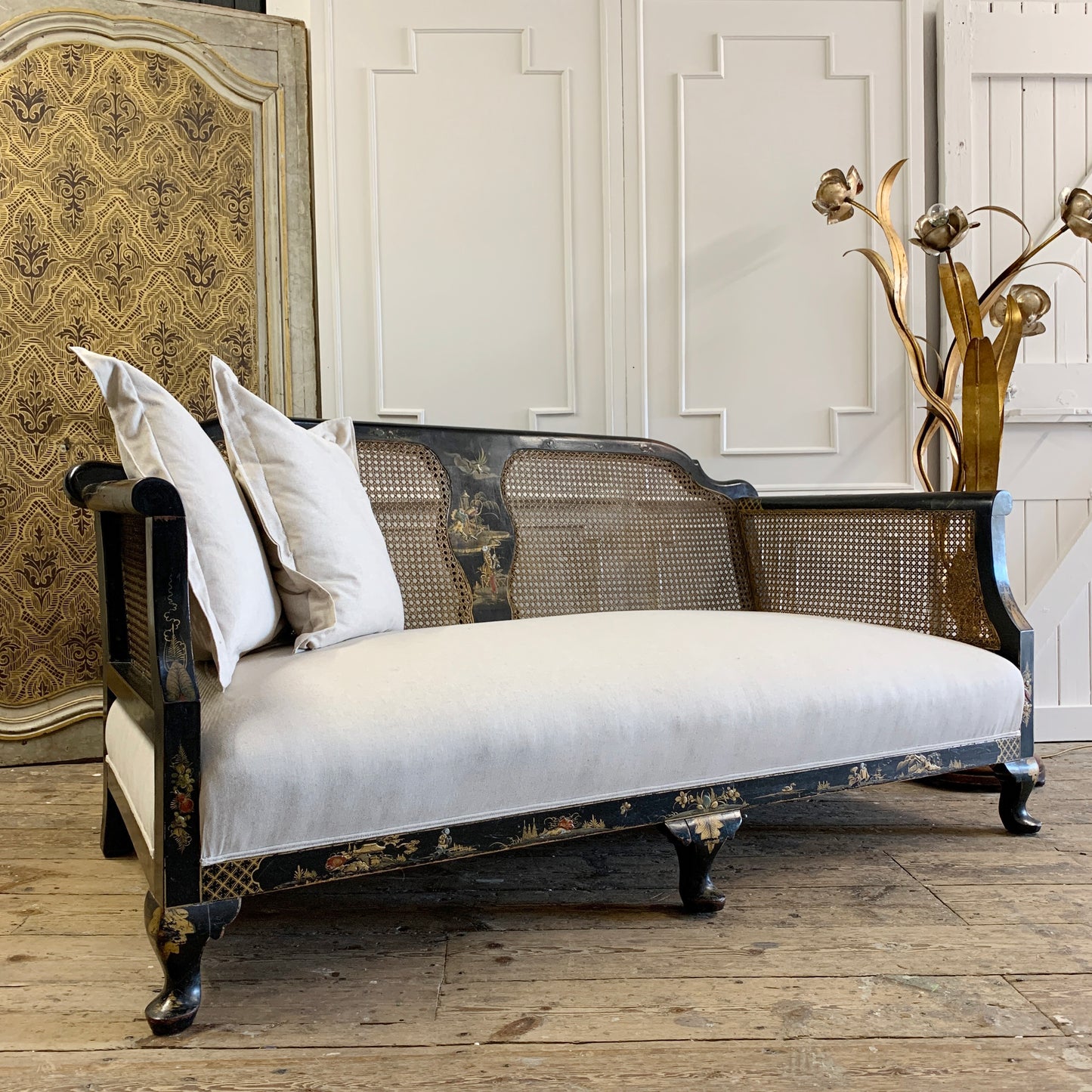 An antique settee with a black and gold chinoiserie lacquered frame, woven cane backrest, and off-white upholstered seat, styled with three white pillows in a rustic room setting.