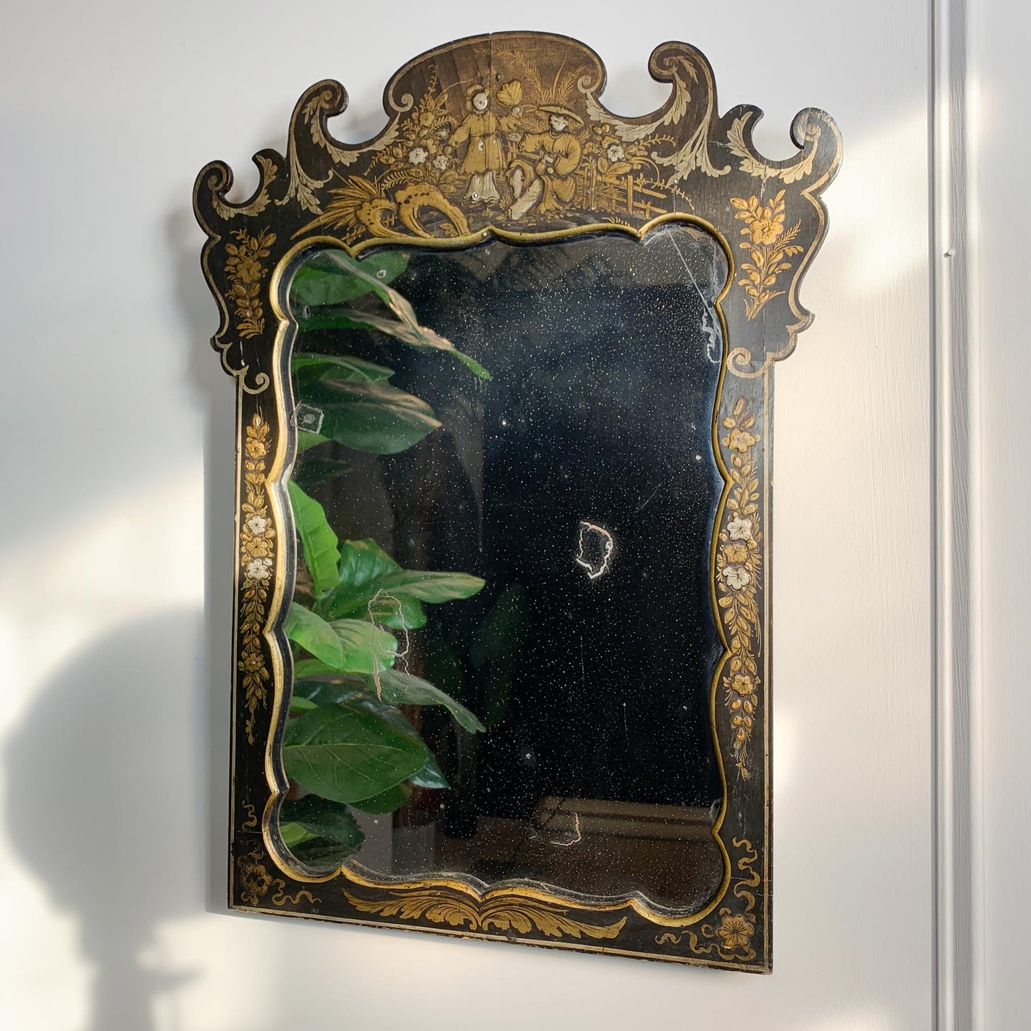 An ornate Napoleon III Black Chinoiserie Mirror from the estate of Jules Verne. mounted on a white wall. The mirror surface is dark and reflects a green houseplant, with some visible wear and damage on the glass.