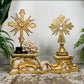 An antique ornate carved wooden religious gold and white carved decorative stand displayed on a  floor against a white wall, with green plants framing the scene and a coral piece between the crosses.