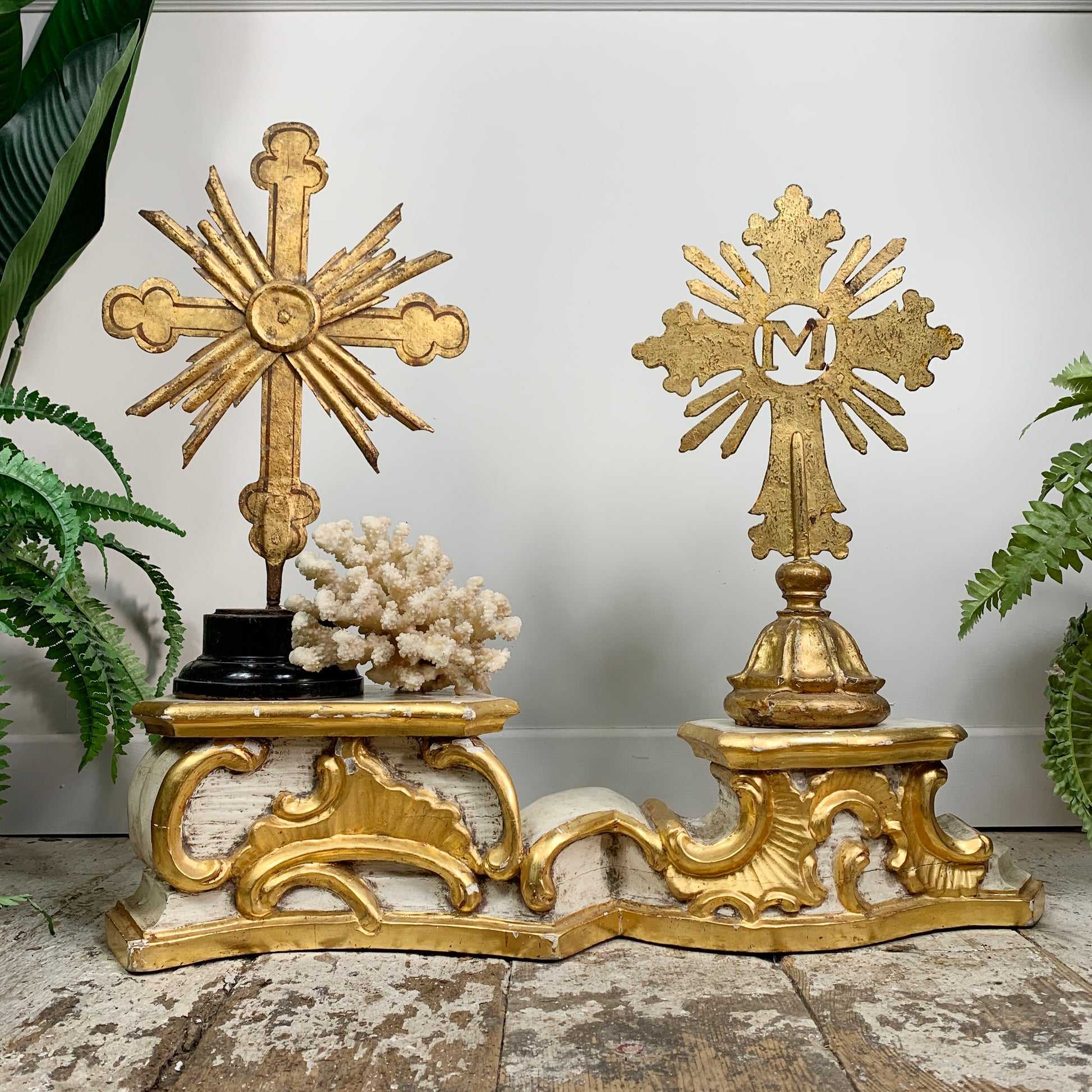 An antique ornate carved wooden religious gold and white carved decorative stand displayed on a  floor against a white wall, with green plants framing the scene and a coral piece between the crosses.