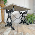 A rustic farmhouse-style table with a light weathered wood top and ornate black cast-iron legs, set on a distressed wooden floor with potted plants in the background.