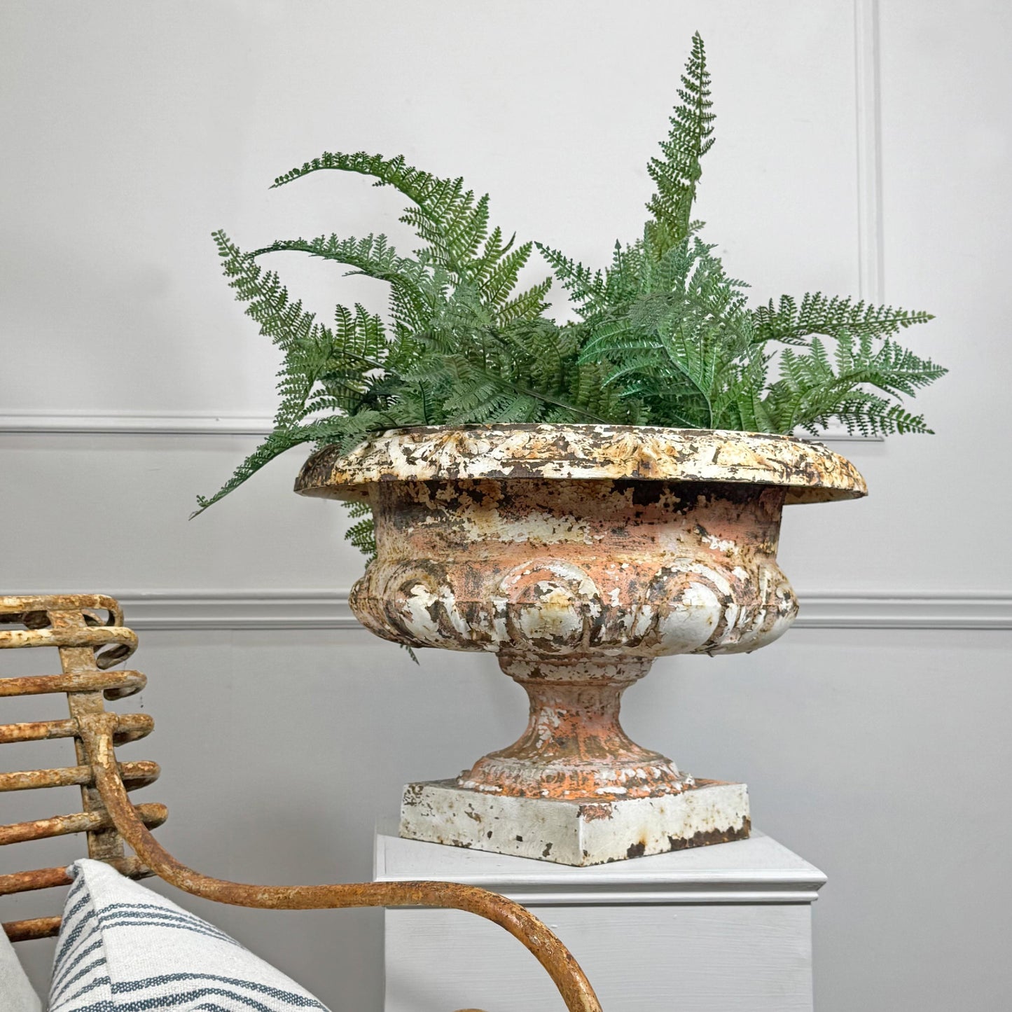 An ornate distressed antique french cast iron urn planter with a weathered white and terracotta finish, filled with green ferns, displayed on a white pedestal in a styled interior setting.