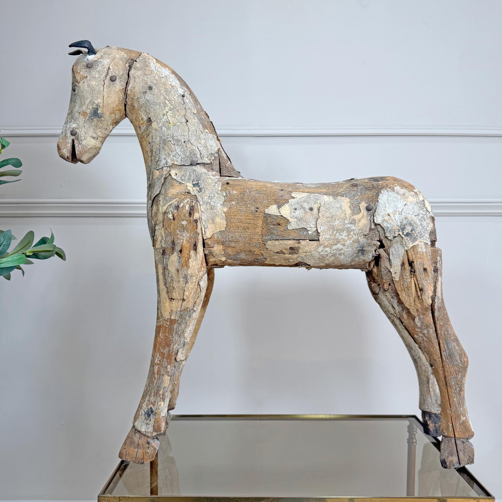 An antique carved wooden horse figurine with a weathered, peeling paint finish, displayed on a mirrored table against a white paneled wall.