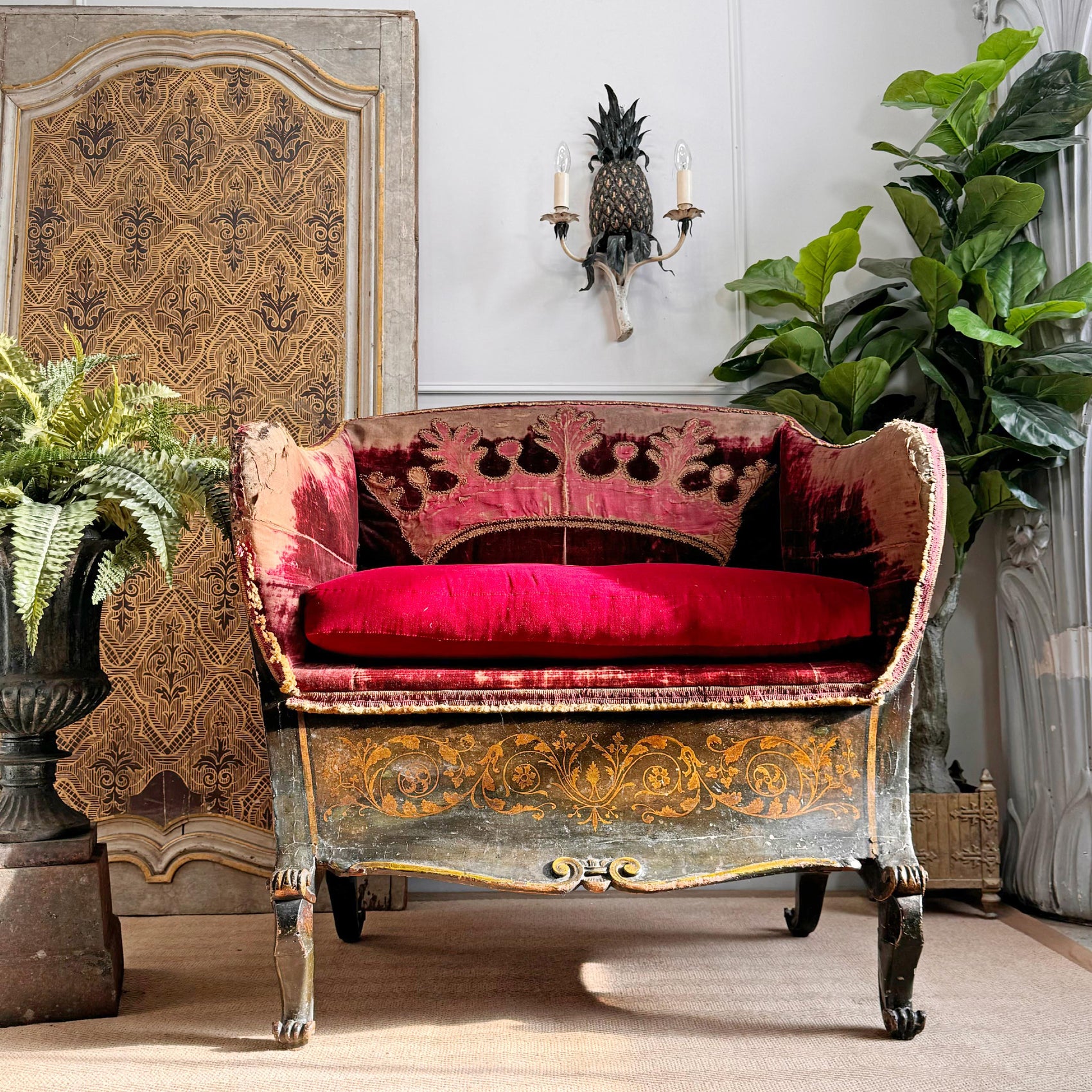 An ornate antique upholstered chair with high detailing, featuring a deep red velvet cushion and burgundy patterned upholstery, styled in a richly decorated room with plants and a decorative screen.