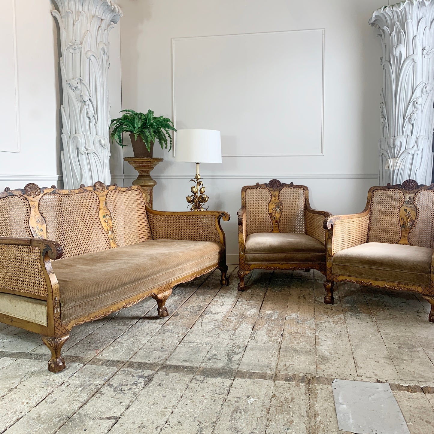 An antique Chinoiserie bergere three-piece furniture set featuring an ornate carved, hand painted gold wooden sofa, two matching armchairs, and a small side table with a lamp, set in a classic room with stone floors and white columns.
