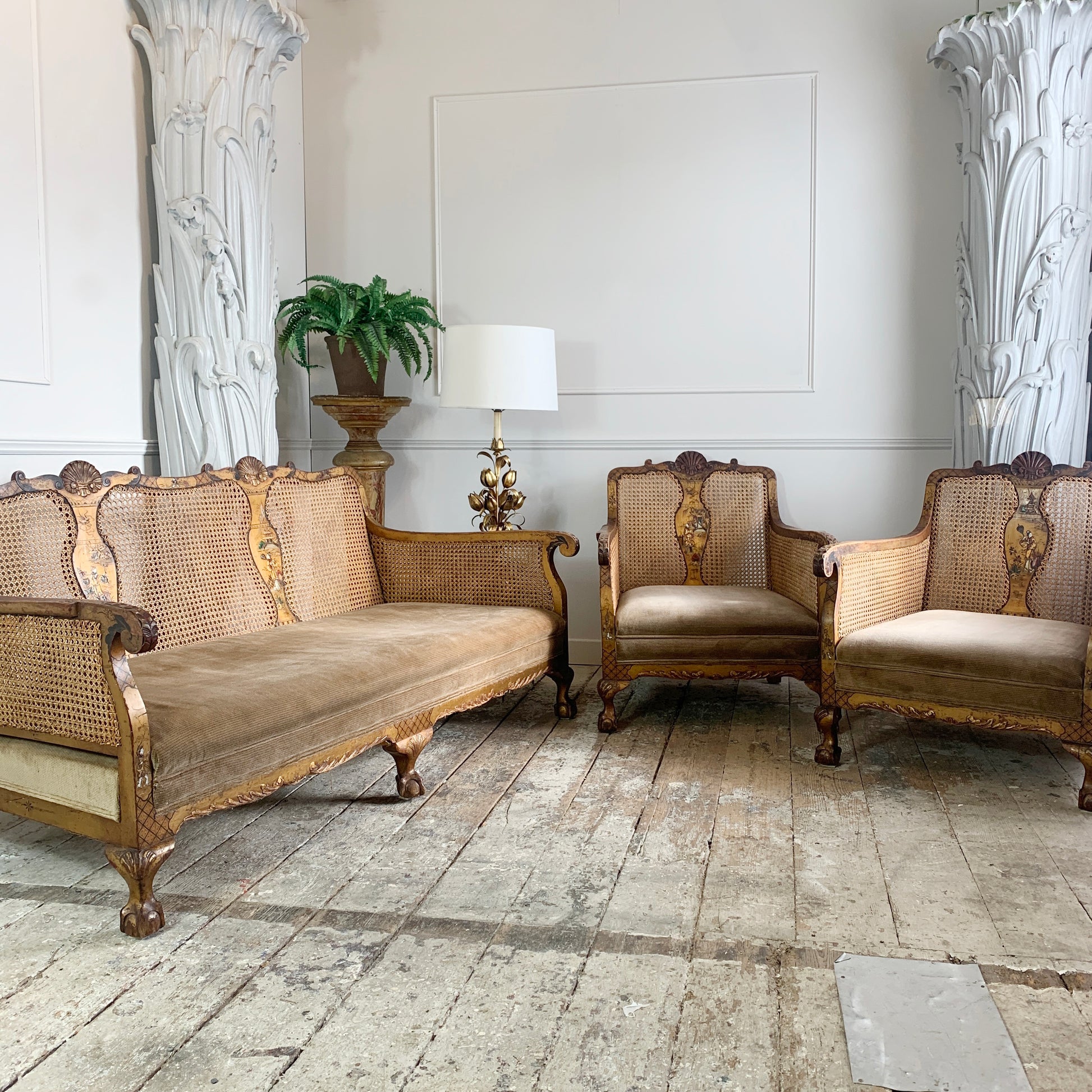 An antique Chinoiserie bergere three-piece furniture set featuring an ornate carved, hand painted gold wooden sofa, two matching armchairs, and a small side table with a lamp, set in a classic room with stone floors and white columns.