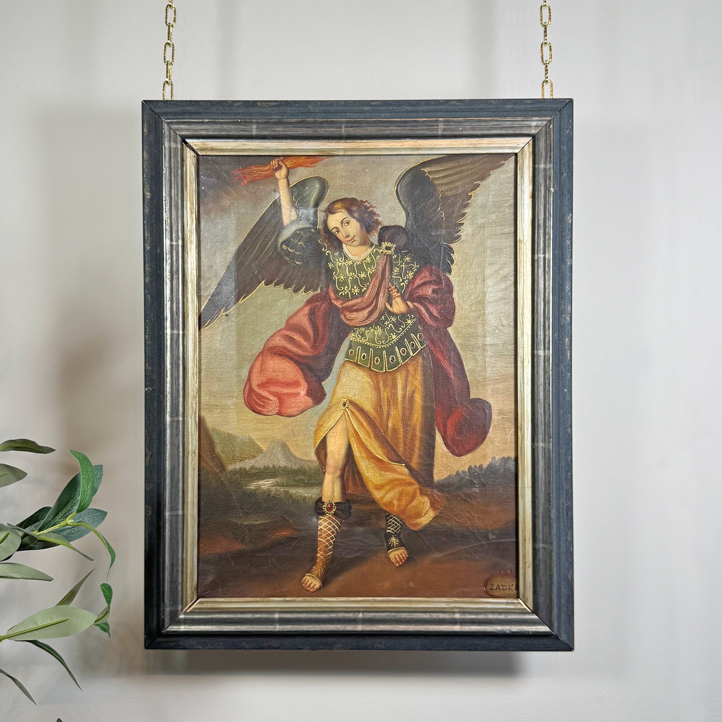 A framed religious Cuzco painting of a winged angel Zadkiel with outstretched wings, holding a sword and shield, hanging on a light wall with a small plant leaf visible at the lower left.