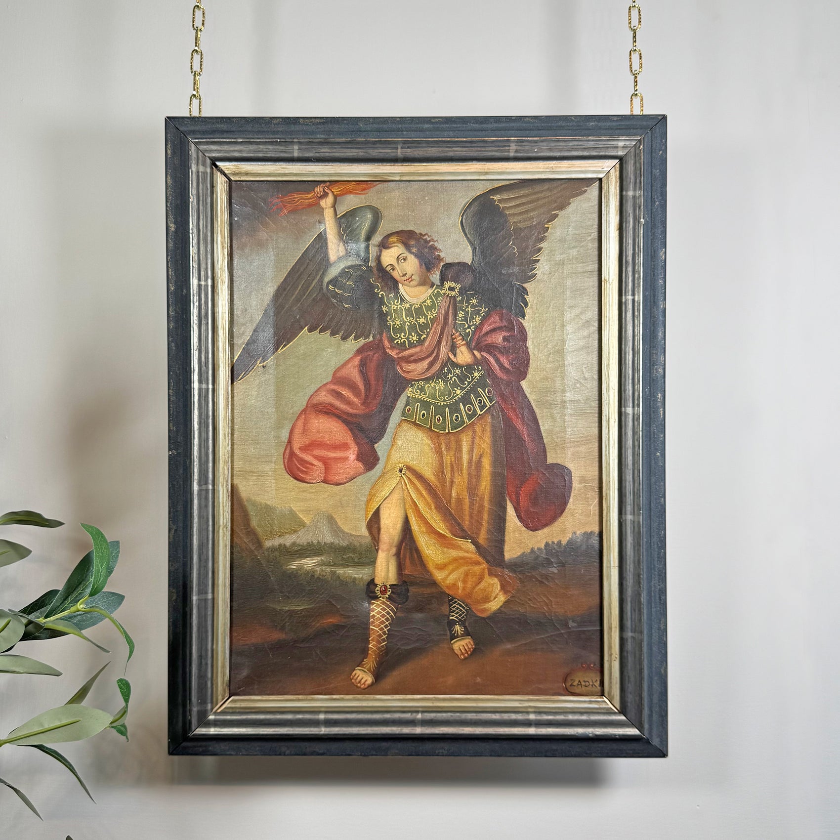 A framed religious Cuzco painting of a winged angel Zadkiel with outstretched wings, holding a sword and shield, hanging on a light wall with a small plant leaf visible at the lower left.