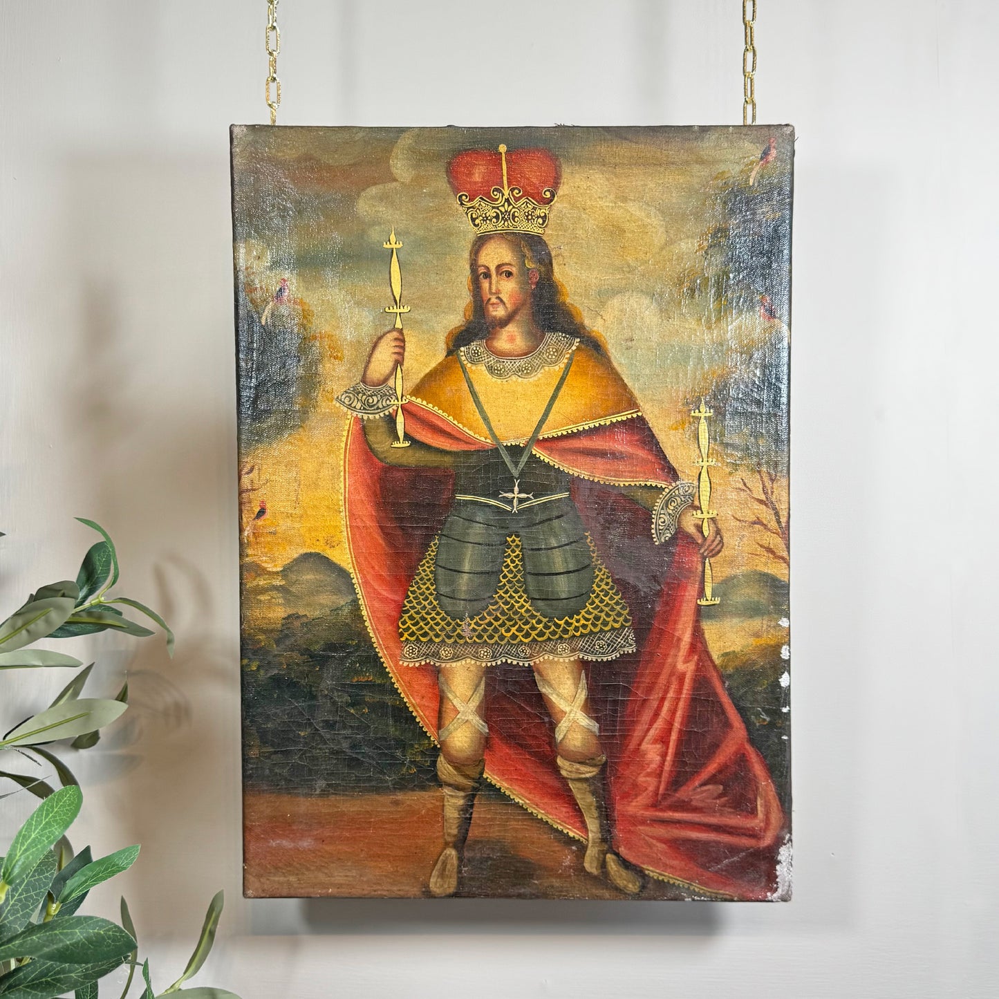 A vertical Cuzco Peruvian painting depicting a crowned king in medieval-style armour with a red cape, hanging on a white wall by two gold chains. A green plant is visible in the lower-left corner.
