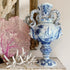 A blue and white antique delft porcelain urn-style vase with sculpted handles, displayed on a shelf next to a pink coral piece and white coral branches.