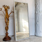 A tall rectangular tailors floor mirror with a distressed white frame leans against a white paneled wall on a light wood floor. To the left is a decorative gold-toned floral table lamp, and to the right is a large white sculptural column.