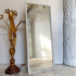 A tall rectangular tailors floor mirror with a distressed white frame leans against a white paneled wall on a light wood floor. To the left is a decorative gold-toned floral table lamp, and to the right is a large white sculptural column.