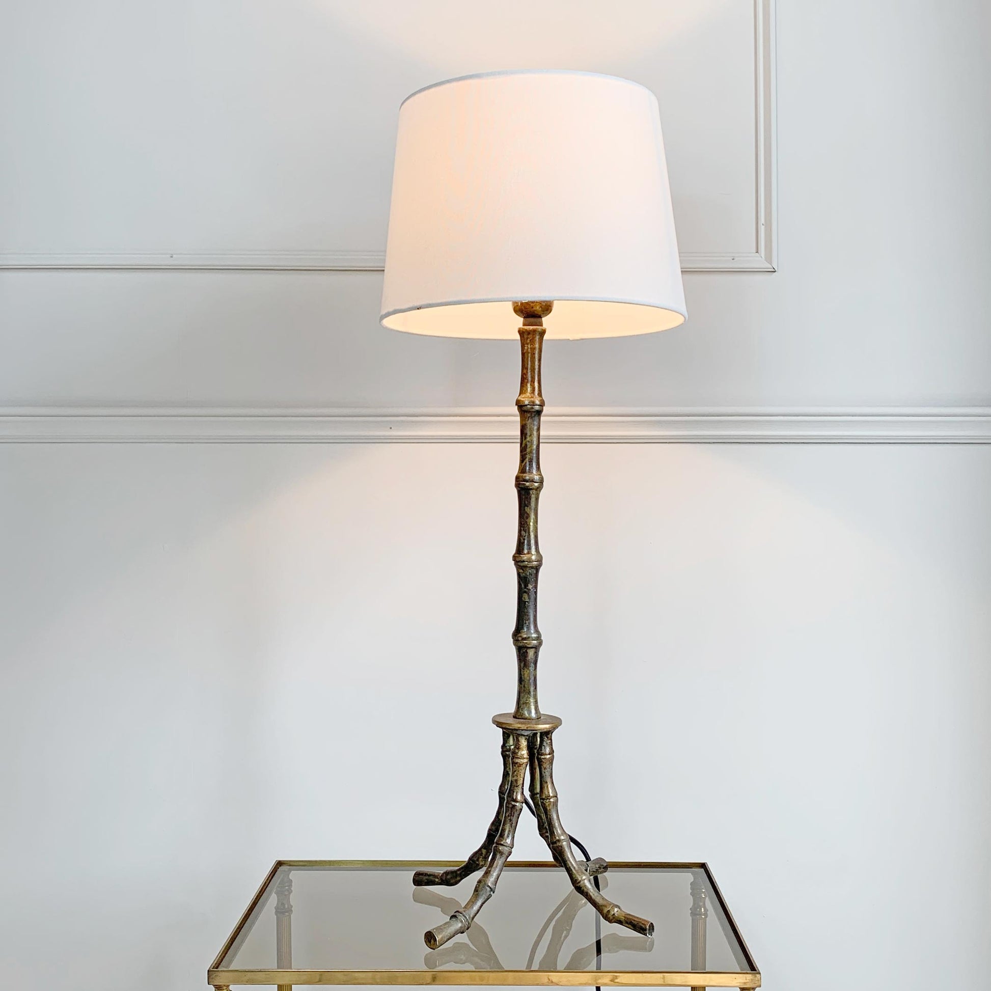 Mid Century Brass Faux Bamboo Table Lamp in the manner of Maison Bagues France, shown lit on a glass-topped side table against a white paneled wall.