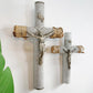 Two antique decorative crucifix wall pieces hanging on a white wall, each with a silver-toned Jesus figure mounted on a cylindrical post wrapped in textured gray rope and a rough wood cross section bound with twine. A green leaf appears at the left edge.