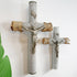 Two antique decorative crucifix wall pieces hanging on a white wall, each with a silver-toned Jesus figure mounted on a cylindrical post wrapped in textured gray rope and a rough wood cross section bound with twine. A green leaf appears at the left edge.