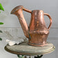 A hammered antique French copper watering can with a long spout and side handle, displayed on a round stone-topped table next to a white decorative object against a light gray wall.