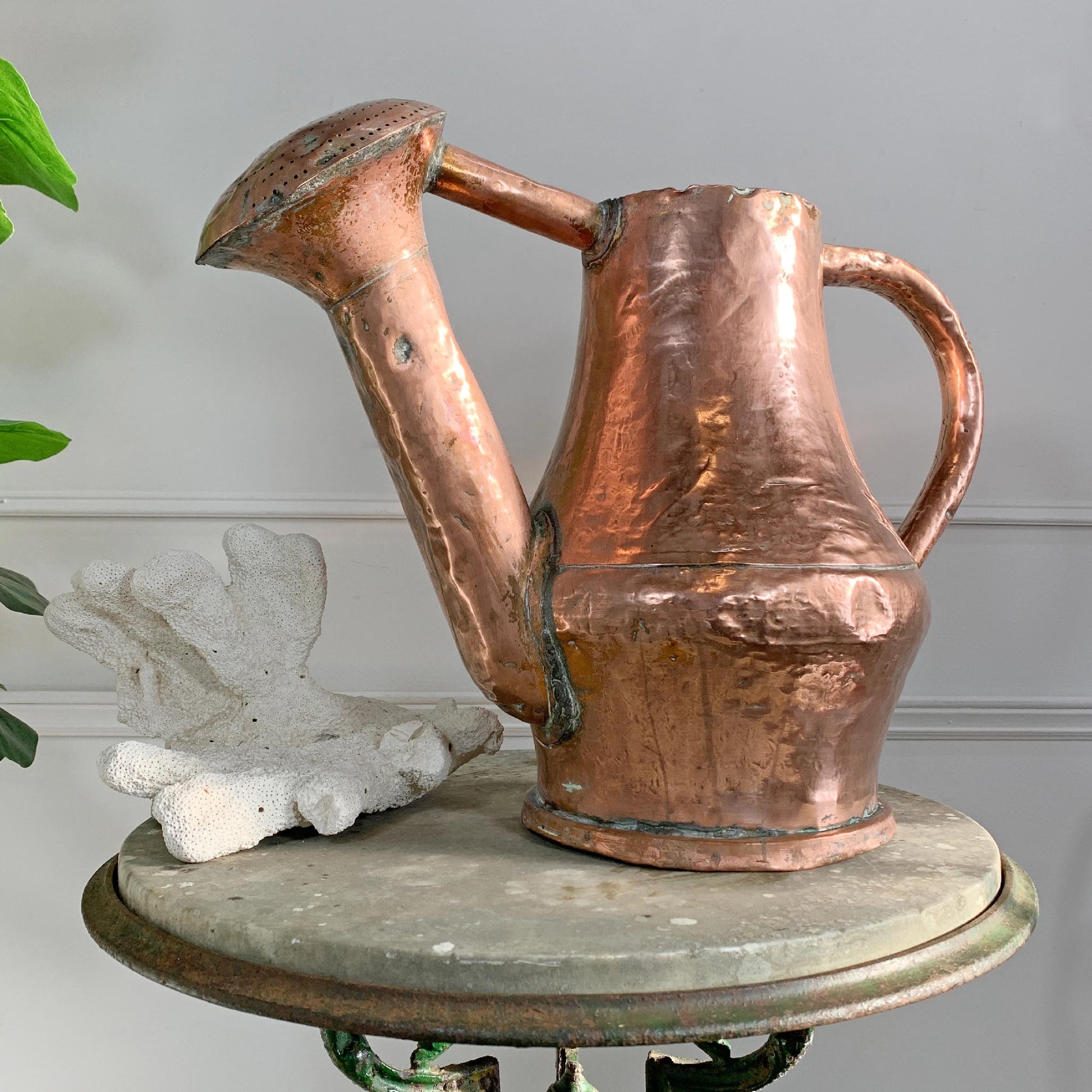 A hammered antique French copper watering can with a long spout and side handle, displayed on a round stone-topped table next to a white decorative object against a light gray wall.