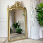 An ornate antique French gold-framed floor mirror leaning against a white panelled wall, with its reflection showing potted plants and a rustic wood floor. A tall white sculptural vase and green houseplants are positioned to the right.