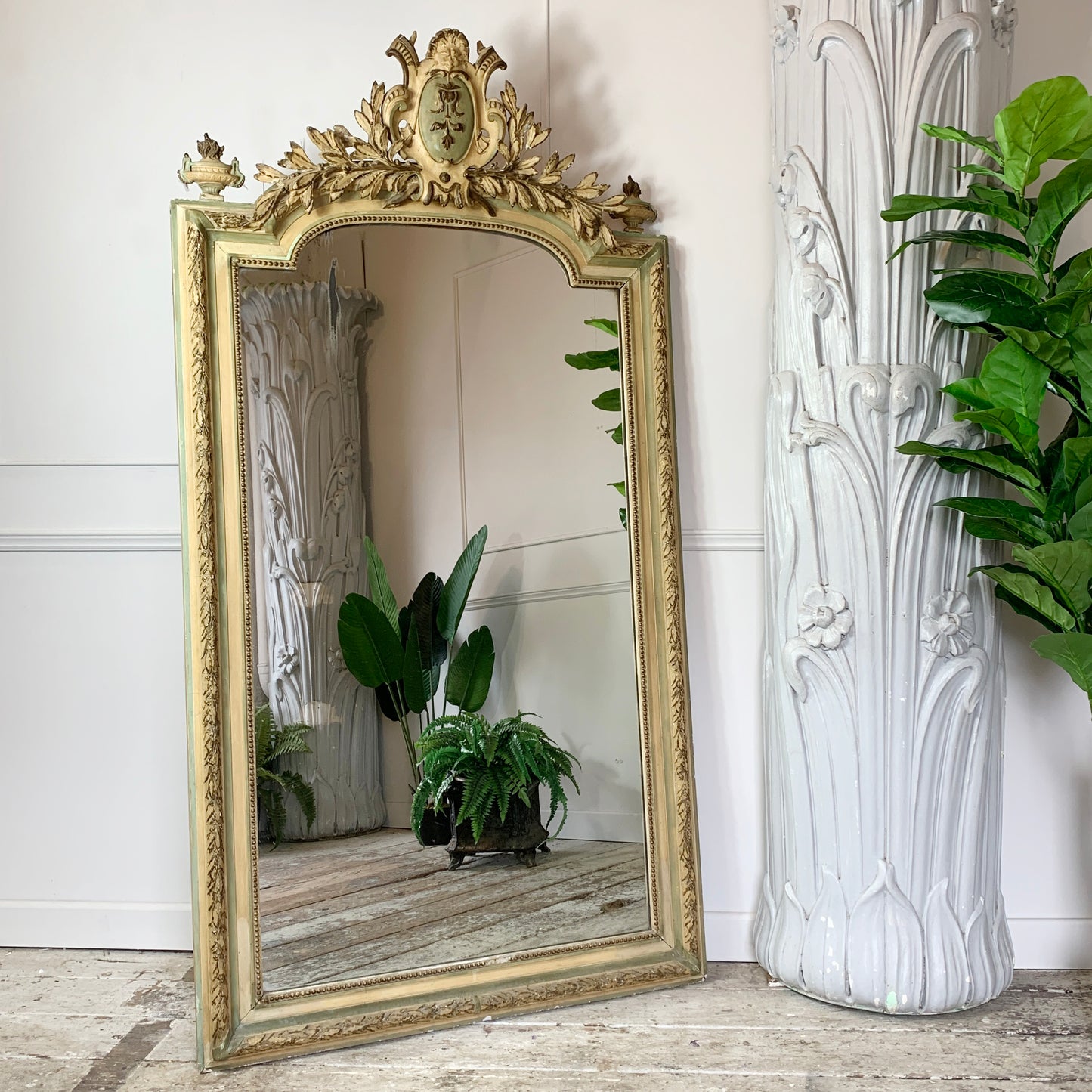 An ornate antique French gold-framed floor mirror leaning against a white panelled wall, with its reflection showing potted plants and a rustic wood floor. A tall white sculptural vase and green houseplants are positioned to the right.