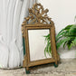 An ornate antique Louis XVI rectangular mirror with a carved gold-toned frame, decorative leaf and branch crest at the top, and small green feet, shown leaning against a white wall with a white sculptural vase and green ferns nearby.