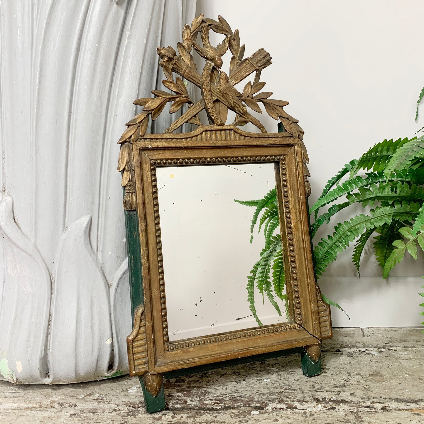 An ornate antique Louis XVI rectangular mirror with a carved gold-toned frame, decorative leaf and branch crest at the top, and small green feet, shown leaning against a white wall with a white sculptural vase and green ferns nearby.