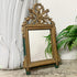 An ornate antique Louis XVI rectangular mirror with a carved gold-toned frame, decorative leaf and branch crest at the top, and small green feet, shown leaning against a white wall with a white sculptural vase and green ferns nearby.