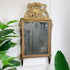 An ornate antique wall mirror with a gilded gold frame, decorative crest at the top, and dark legs, shown leaning against a white wall with green houseplants partially visible on both sides.