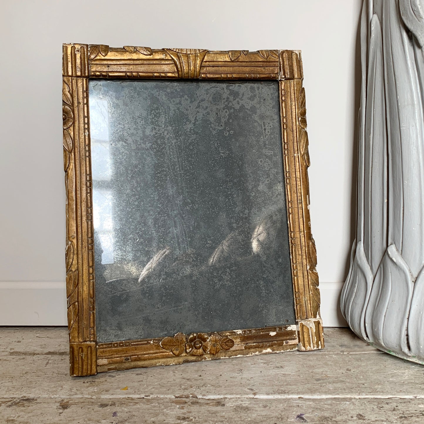 An ornate antique rectangular mirror with a distressed gold frame, leaning against a white wall on a rustic wooden floor next to a gray decorative pillar.