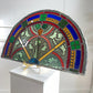 A large semi-circular stained glass panel with ornate floral and scroll motifs, displayed on a white pedestal. The design features blue, red, green, yellow, and white glass sections held together by dark metal lead lines.
