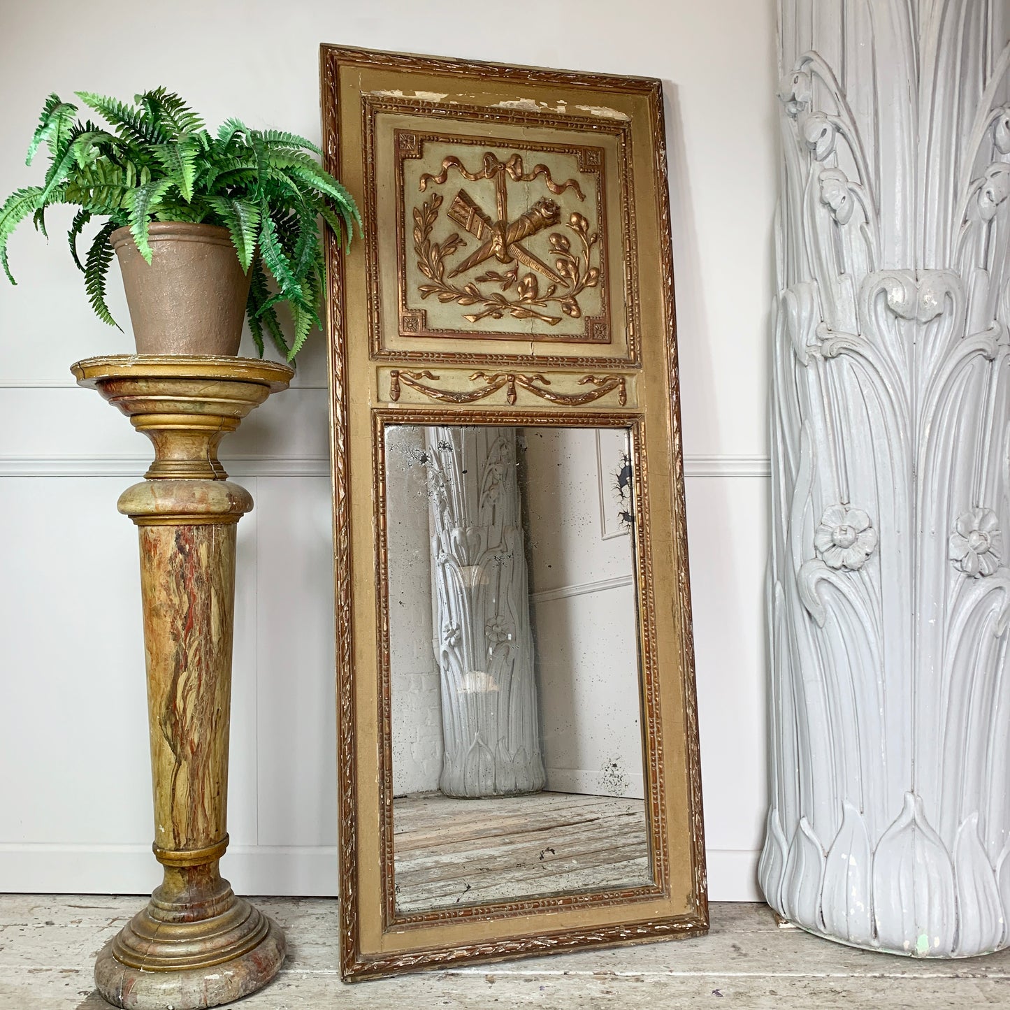 An ornate antique full-length mirror with a distressed gold and beige carved frame, leaning against a white wall on a light wood floor. A potted green fern sits on a matching gold pedestal to the left, and a large white sculptural pillar is on the right.