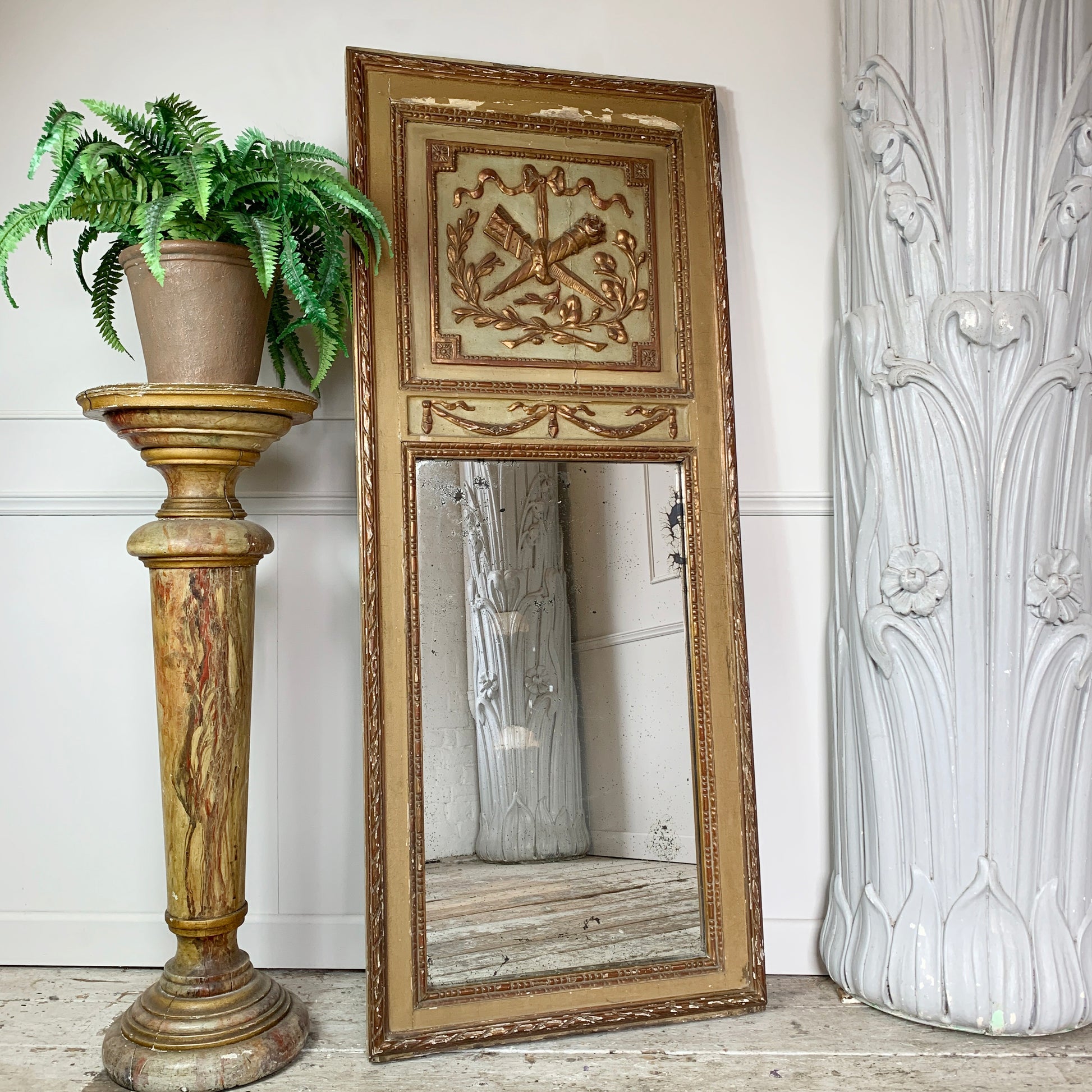 An ornate antique full-length mirror with a distressed gold and beige carved frame, leaning against a white wall on a light wood floor. A potted green fern sits on a matching gold pedestal to the left, and a large white sculptural pillar is on the right.