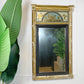 An ornate rectangular regency wall mirror with a distressed gold frame and a decorative painted crest panel at the top, shown hanging on a white wall next to a large green plant.