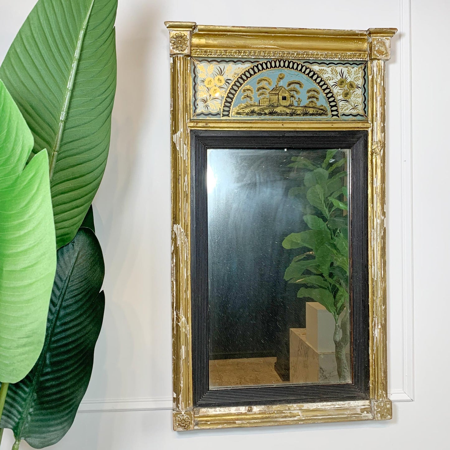 An ornate rectangular regency wall mirror with a distressed gold frame and a decorative painted crest panel at the top, shown hanging on a white wall next to a large green plant.