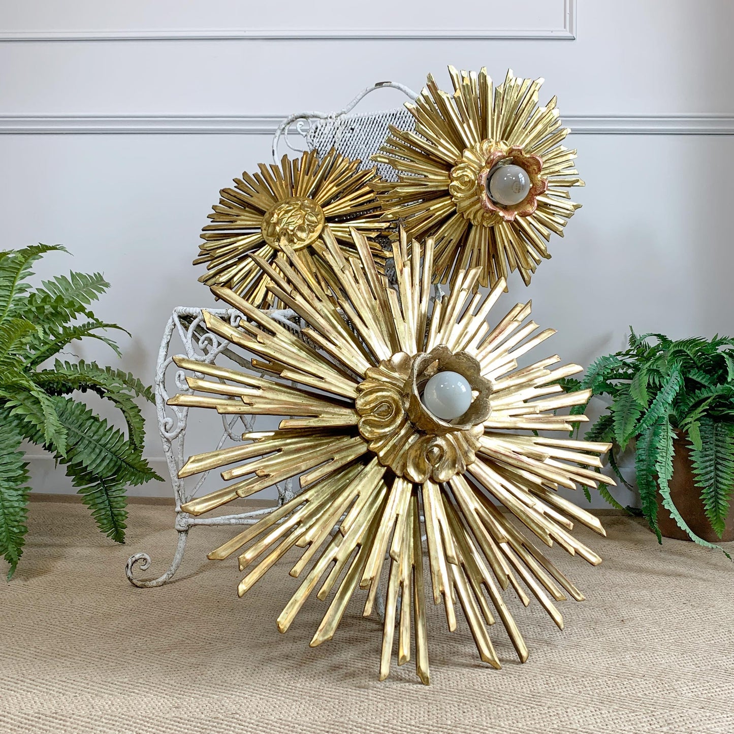 Gold sunburst wood and gesso wall decor with three flower-like spheres and central light bulbs, styled on a beige surface with potted ferns and a white chair in the background.