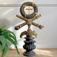 A decorative antique religious sculpture featuring a wreath with a cross, crossed swords, a celtic-style crest, and a dark pedestal base, styled indoors next to a green fern plant.